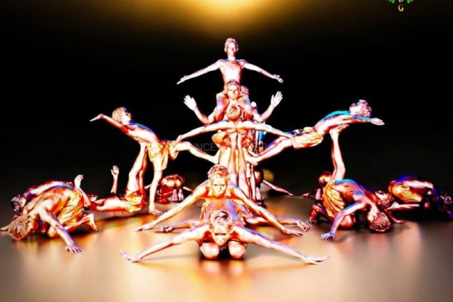 Prince Dance Group performers in gold body paint forming an acrobatic pyramid with arms extended, under warm stage lighting